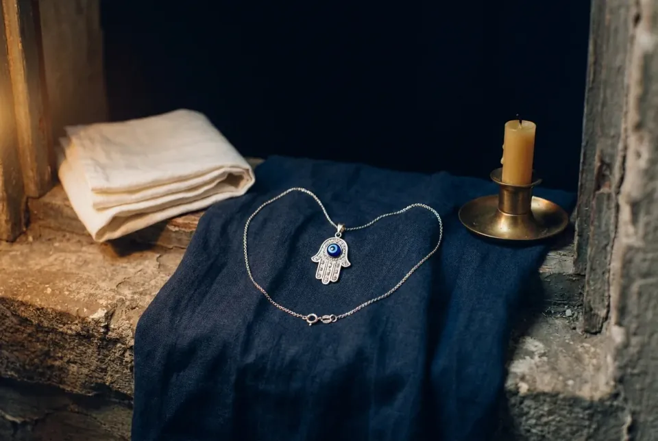 Silver hamsa pendant with a blue-and-white eye on indigo linen beside folded cloth and an unlit candle
