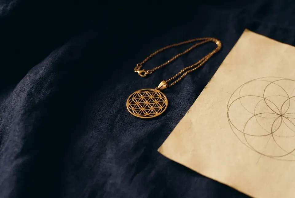Gold Flower of Life pendant on dark linen beside an unfinished overlapping-circle drawing